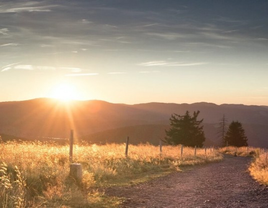 Sicht von einem Feldweg auf einen beschaulichen Sonneruntergang hinter einem Bergpanorama.