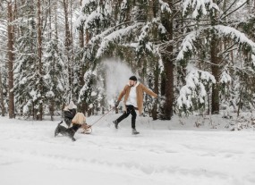 Ein Mann zieht laufend im Schnee eine Person auf einem Schlitten hinter sich her und wird von einem Hund begleitet.